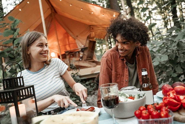 日没後に笑いながら、グランピングで夕食をとる若い多民族のカップル。電球のライトの下で野外ピクニックでキャンプする幸せなミレニアル世代。アウトドアで友達と過ごす時間、バーベキューパーティー