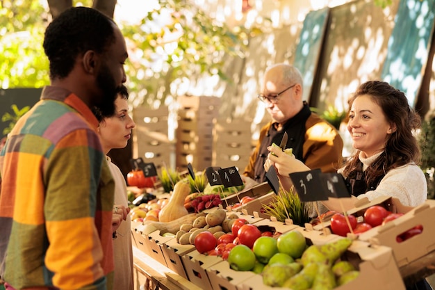 Photo young multiracial couple man and woman buying fresh organic produce at farmers market. smiling local female vendor standing behind fruit and vegetable stand offering customers to try apple