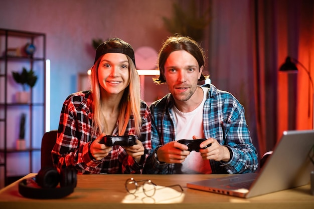young multiracial couple sitting at table with wireless joysticks in hands and playing video games
