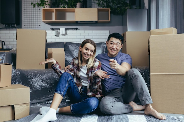 Young multiracial family man and woman sitting on the floor among cardboard boxes looking at the camera and smiling waving hands greeting gesture