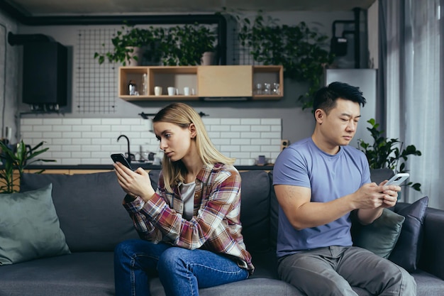 Young multiracial family quarreling sitting on the couch in the room man and woman on the phone turned in different directions