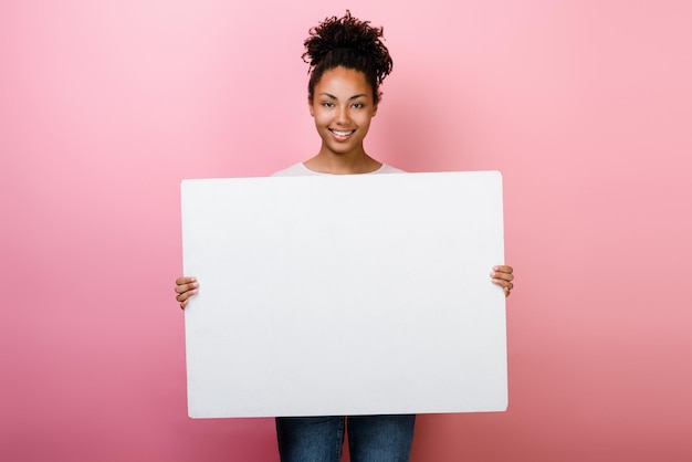Молодая многорасовая женщина показывает и держит пустой белый рекламный щит на розовом фоне Рекламная концепция