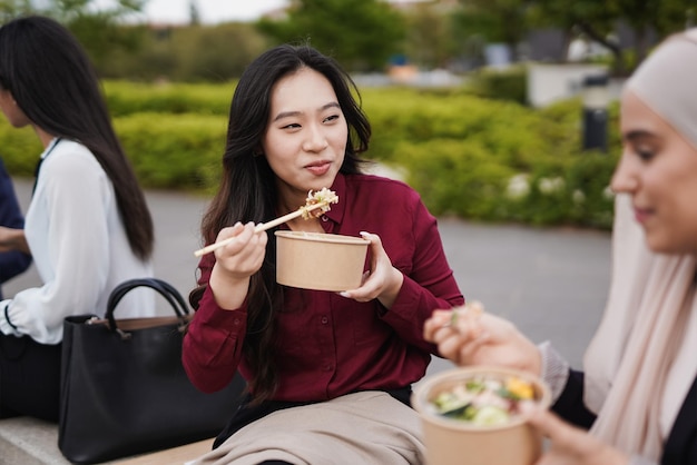 Young multiracial women eating together healthy poke food\
during lunch break outside of office