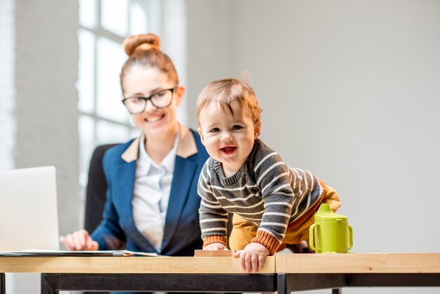 Young multitasking businessmam dressed in the suit working with laptop and documents sitting with her baby son at the office