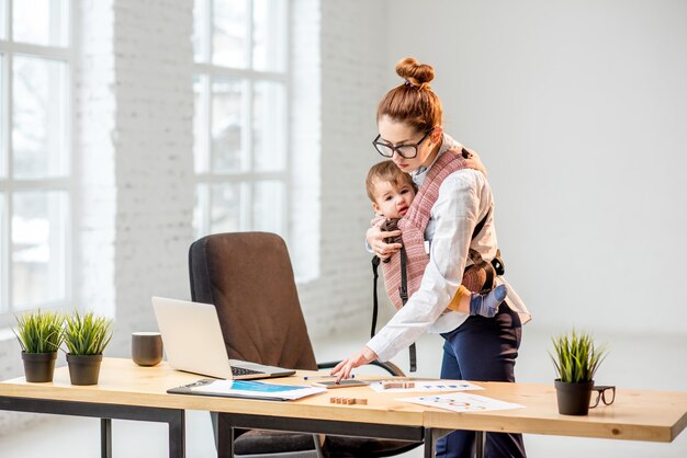 Young multitasking businesswoman standing with her baby son during the work at the office