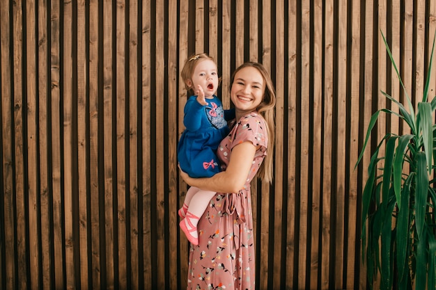 Young mum holds her daughter on hands over wooden wall