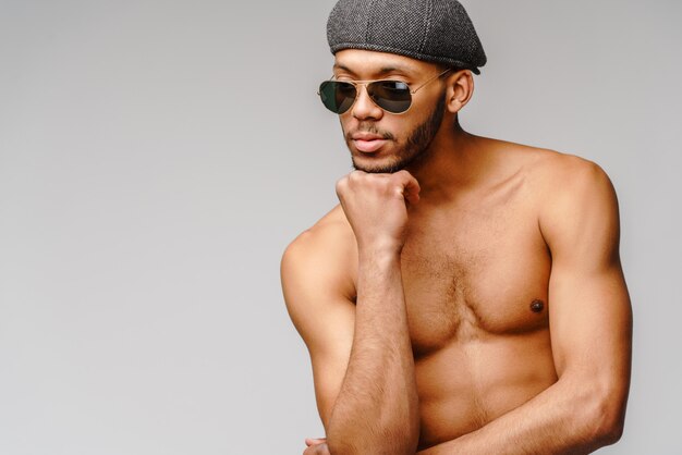  young muscular african american man shirtless wearing sunglasses and cap over light grey wall