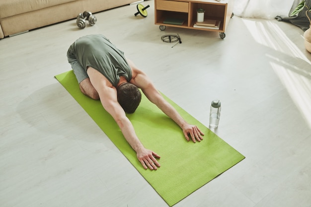 Молодой мускулистый спортсмен делает упражнения на расслабление на коврике, стоя на коленях с вытянутыми вперед руками во время домашней тренировки