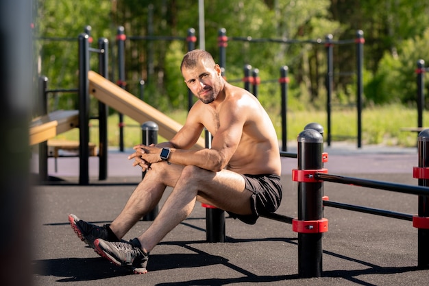 Young muscular athlete sitting on bar of sports facilities outdoors while having break between trainings