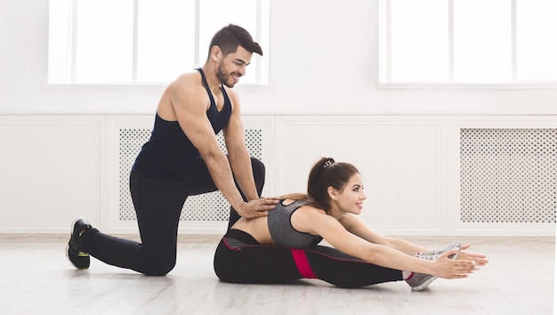 Young muscular coach helping girl doing stretching exercise in light studio, panorama, free space