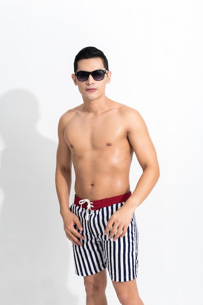 Young muscular guy in striped beach shorts sunbathing wear sunglasses at studio shot isolated on white background. Fashion summer concept.