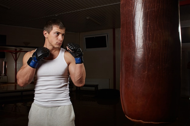 Young muscular man boxer workout with punching bag in dark interior