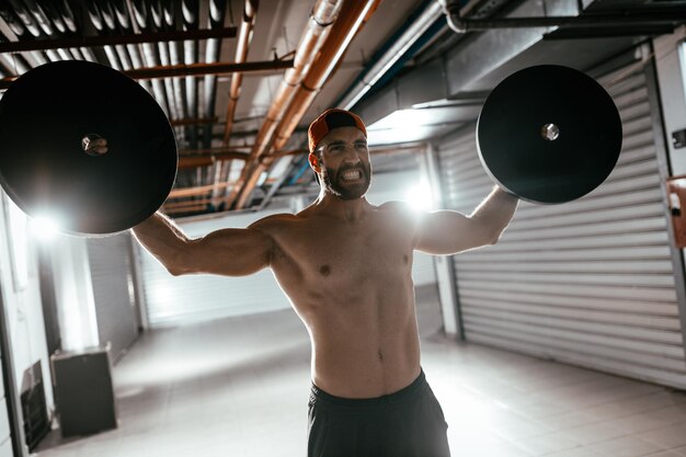 Young muscular man doing hard exercise with weights for shoulders on cross fit training at the garage gym.