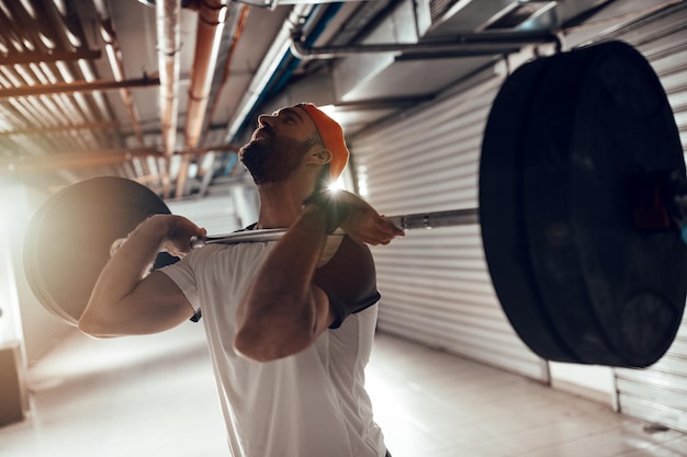 Молодой мускулистый мужчина делает упражнения с высокой тягой со штангой на перекрестных тренировках в гаражном спортзале.