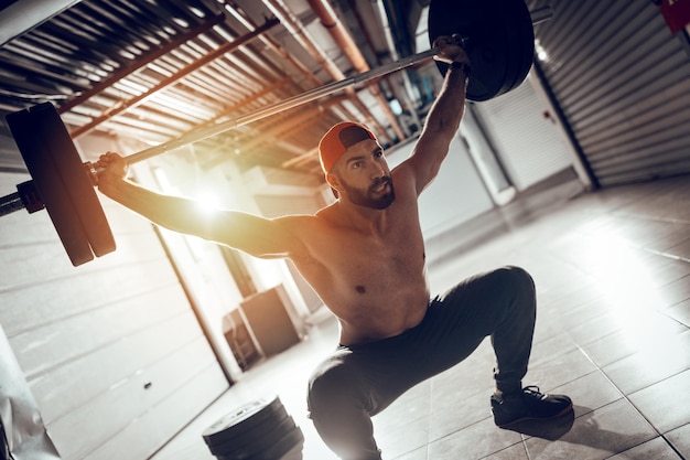 Young muscular man doing overhead squat exercise with barbell on cross training at the garage gym.