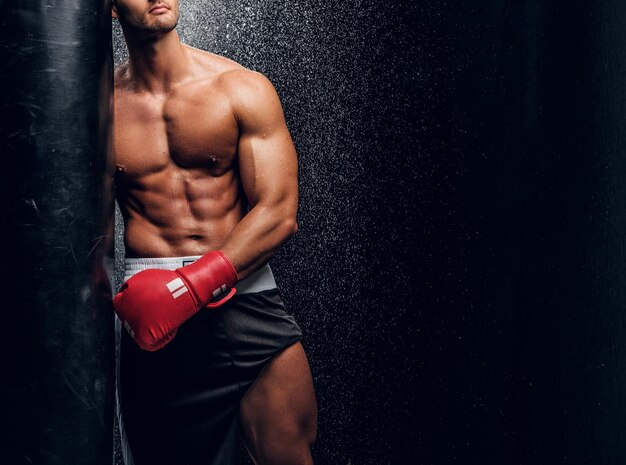 Young muscular man in red boxer gloves is posing with punching bag at dark room with water splashes on the wall.