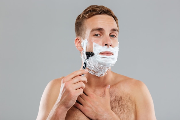 Young muscular man shaving over gray wall and looking at front