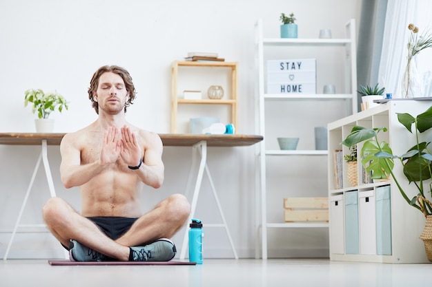 Young muscular man sitting on exercise mat with his eyes closed and doing breathing exercises at home