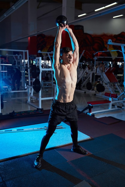 Young muscular man with naked torso working out in gym Athletic male adult exercising with kettle bell Fitness sports concept