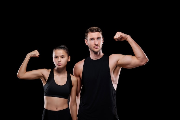 Young muscular sportsman and pretty fit female in activewear showing their strength while standing