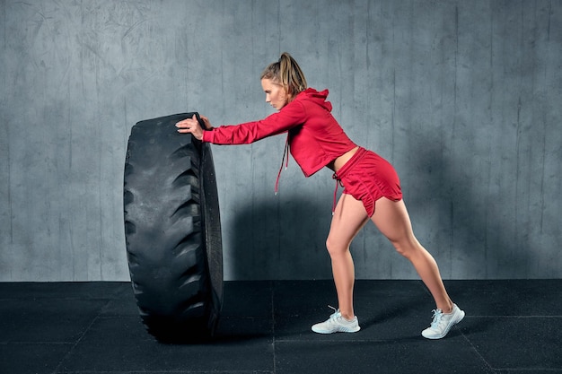Молодая мускулистая женщина переворачивает шину на тяжелых тренировках с личным тренером в спортзале гаража.