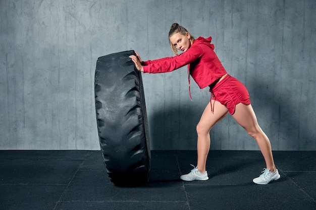 ガレージジムでパーソナルトレーナーとのハードトレーニングでタイヤをひっくり返す若い筋肉の女性。