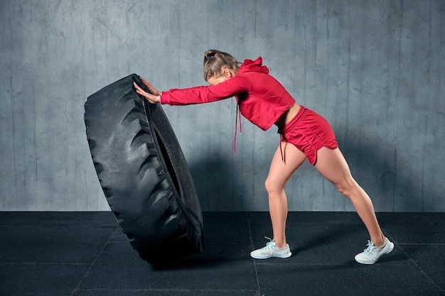 ガレージジムでパーソナルトレーナーとのハードトレーニングでタイヤをひっくり返す若い筋肉の女性。