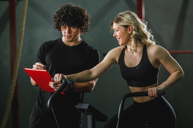 Foto una giovane donna muscolosa sta facendo un allenamento crossfit in bici in palestra supportata da un allenatore.