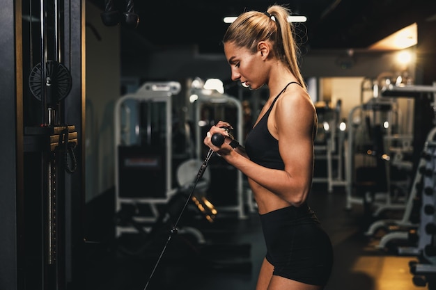 若い筋肉質の女性がジムでマシンのトレーニングを行っています。彼女は重い体重で腕の筋肉をポンプでくみ上げています。