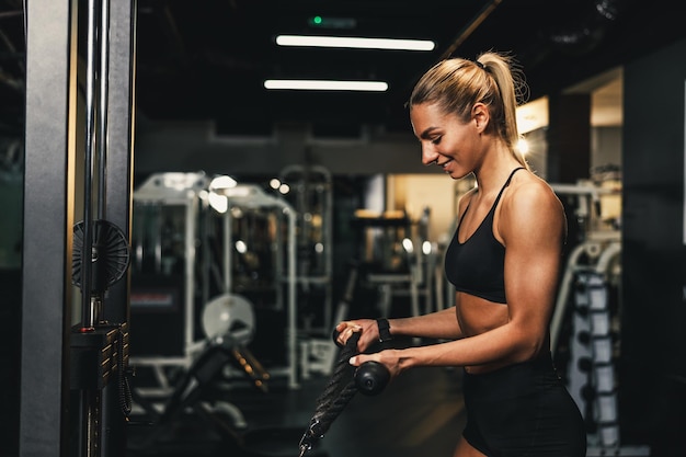 젊은 근육질의 여성이 체육관에서 기계 훈련을 하고 있습니다. 그녀는 무거운 무게로 팔 근육을 펌핑하고 있습니다.