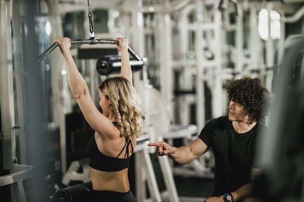 Photo a young muscular woman is doing training at machine supporting by coach in the gym.