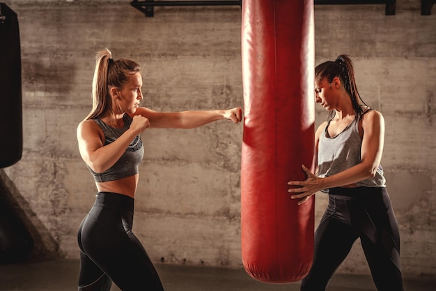 Young muscular woman punching a boxing bag on cross fit training with a female personal trainer at the gym.