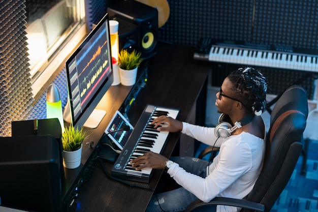 Молодой музыкант африканской национальности сидит в студии записи, создает новую музыку и микширует звуки на компьютере