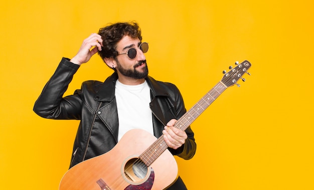 Young musician man feeling puzzled and confused, scratching head and looking to the side with a guitar, rock and roll concept