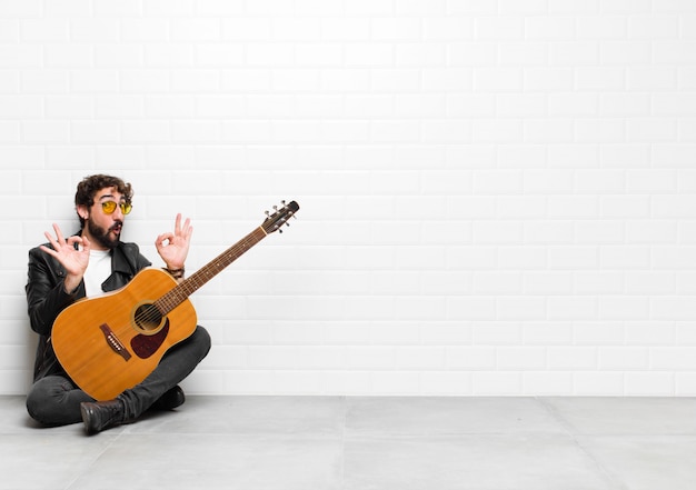 Young musician man feeling shocked, amazed and surprised, showing approval making okay sign with both hands with a guitar, rock and roll concept