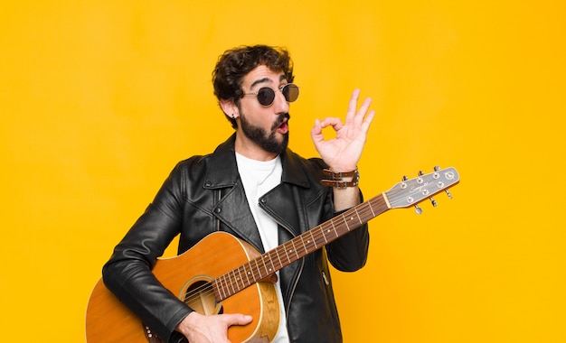 Young musician man feeling successful and satisfied, smiling with mouth wide open, making okay sign with hand with a guitar, rock and roll concept