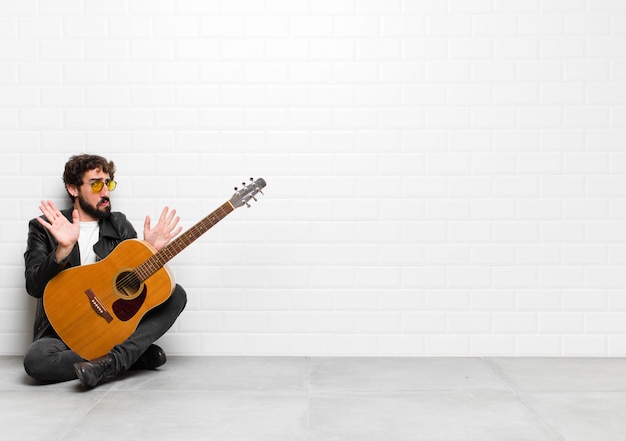 Young musician man looking nervous, anxious and concerned, saying not my fault or I didnÃ¢ÂÂt do it with a guitar, rock and roll concept