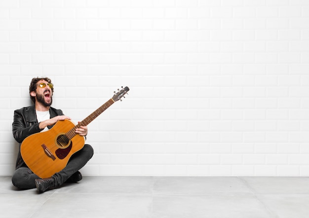 Young musician man screaming furiously, shouting aggressively, looking stressed and angry with a guitar, rock and roll concept