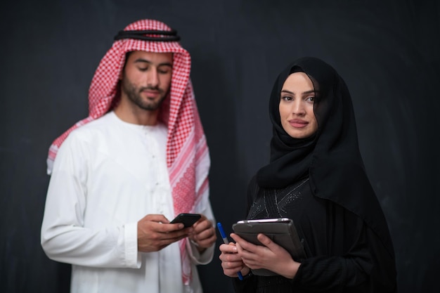 Young muslim business couple arabian man with woman in fashionable hijab dress using mobile phone and tablet computer in front of black chalkboard representing modern islam fashion technology.