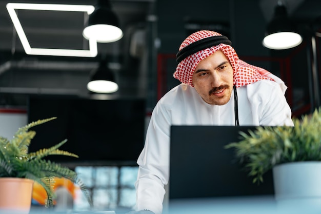 Молодой мусульманский бизнесмен в традиционной одежде работает за столом в офисе