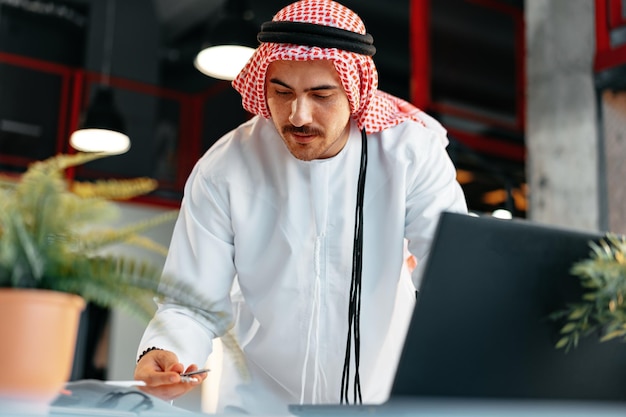 オフィスのテーブルで働く伝統的な服を着た若いイスラム教徒のビジネスマン