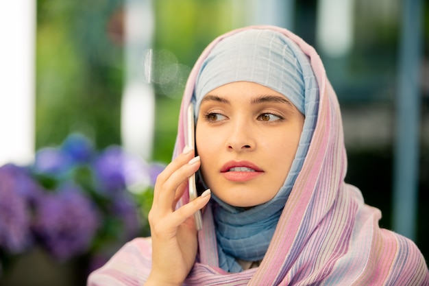 Young muslim businesswoman in hijab talking on smartphone outdoors while consulting client or colleague