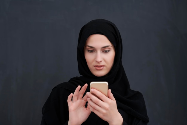 Young muslim businesswoman in traditional clothes or abaya\
using smartphone. arab woman standing in front of black chalkboard\
and representing techology, islamic fashion and ramadan kareem\
concept