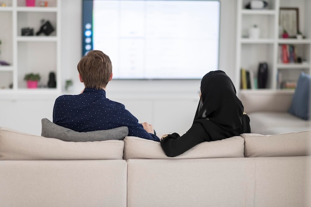 Young Muslim couple woman wearing Islamic hijab clothes sitting on the sofa watching TV together during the month of Ramadan at a modern home.