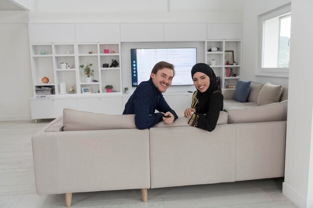 Young Muslim couple woman wearing Islamic hijab clothes sitting on the sofa watching TV together during the month of Ramadan at a modern home.