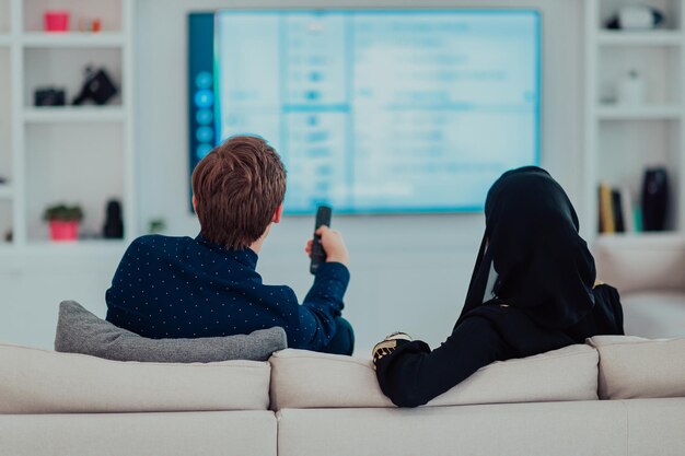 Young muslim couple woman wearing islamic hijab clothes sitting on sofa watching TV together during the month of Ramadan at modern home.