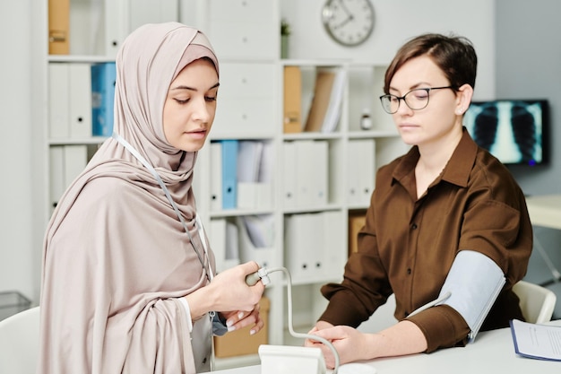 Young muslim female physician checking blood pressure of patient by workplace