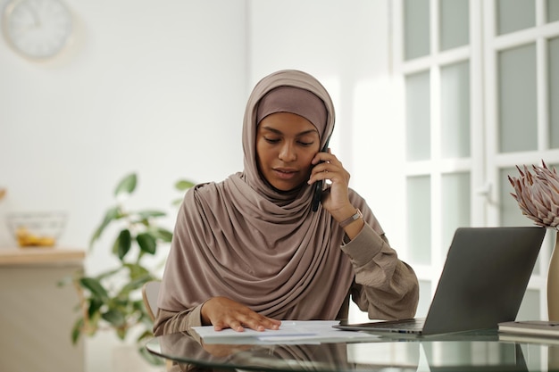 Young Muslim woman in beige headscarf looking at document during call on smartphone