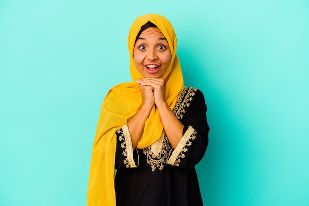 Young muslim woman on blue praying for luck, amazed and opening mouth looking to front.