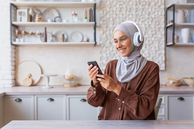 A young muslim woman in a hijab is sitting at home in the kitchen wearing headphones holding a phone